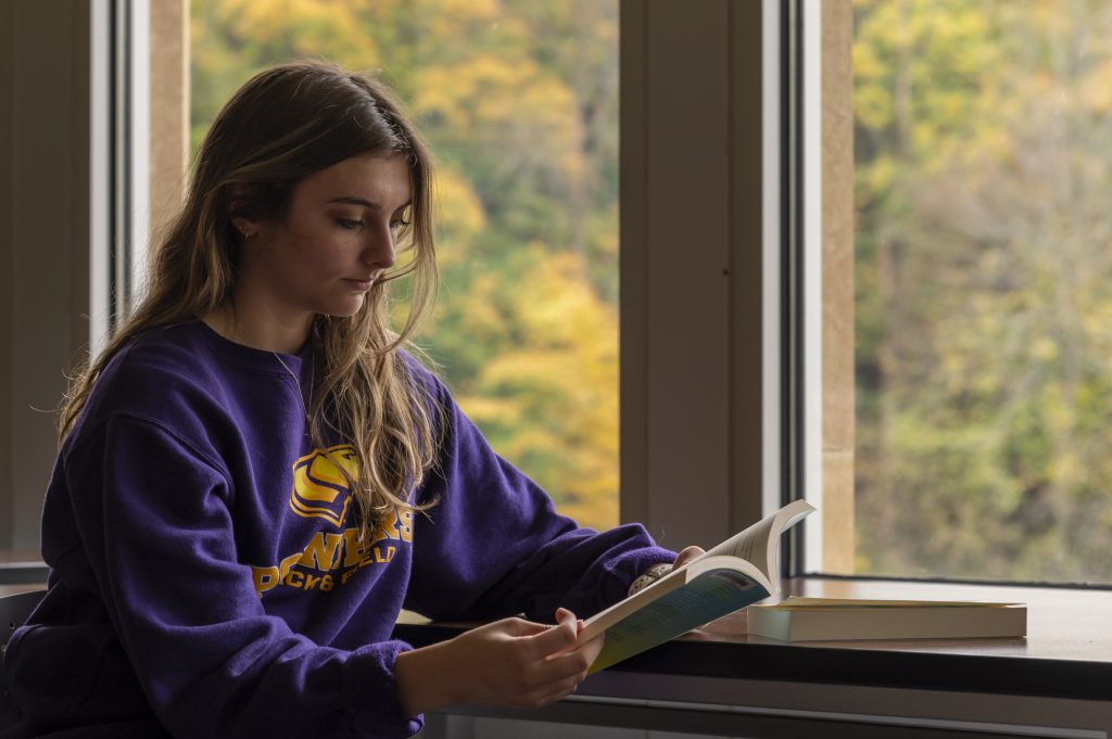 A female student reading in the library in a purple and gold Pointer sweatshirt.