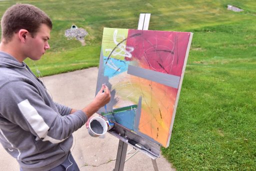 A male student painting an abstract piece outside while listening to music.