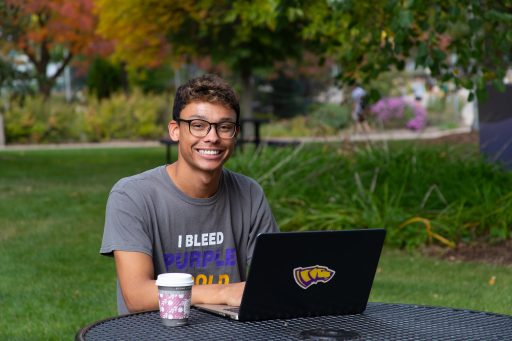 A student at a picnic table smiling and typing on a laptop computer with a coffee next to him.