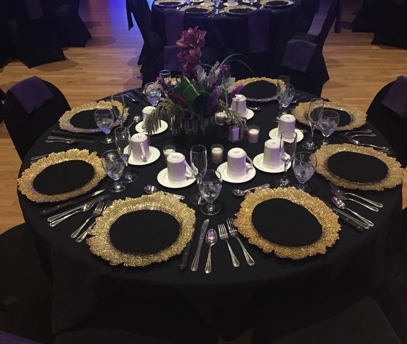 Table setting with black tablecloth and chair covers, gold and black plates and purple flowers
