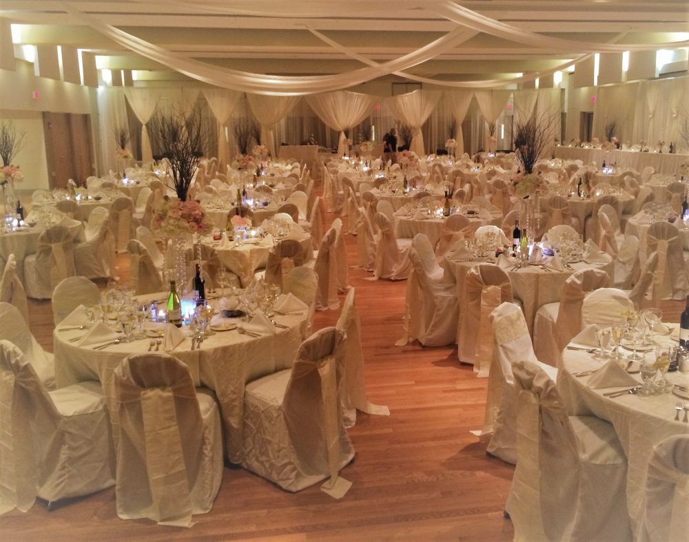 Laird room decorated with white drapes, dinner tables set with white table clothes and chair covers, pink flowers and wine bottles.