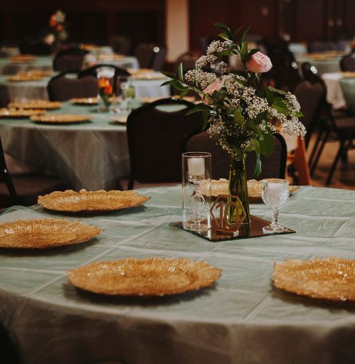 Table setting with gold plates, pink flowers, babies breathe and leaves. Glasses vases with candles sitting on a mirror.