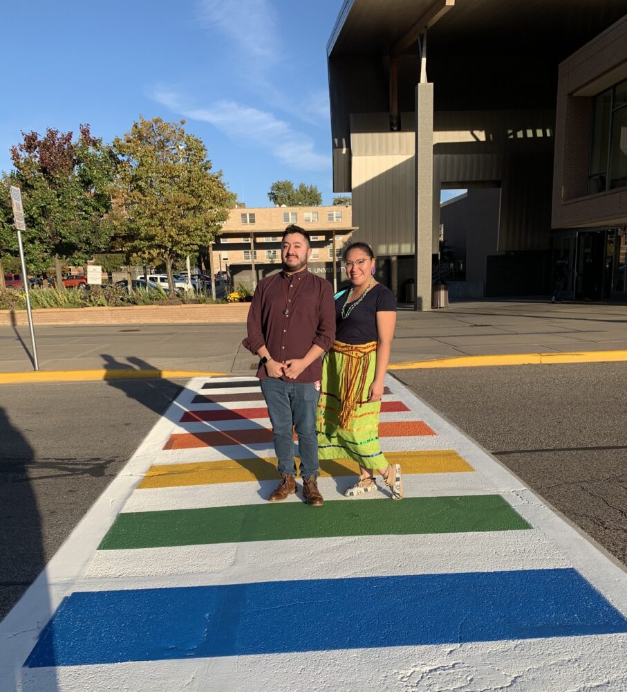 Two people standing on a LGBTQ+ themed crosswalk in front of the Dreyfus University Center.