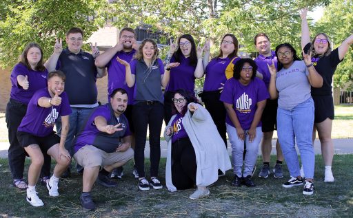 Photo of Hall Director Team. Smiling poses wearing purple shirts. Staff giving thumbs up and peace sign. 