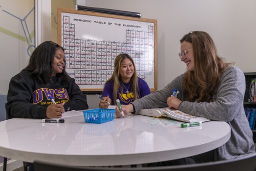 3 students working in front a periodic table whiteboard